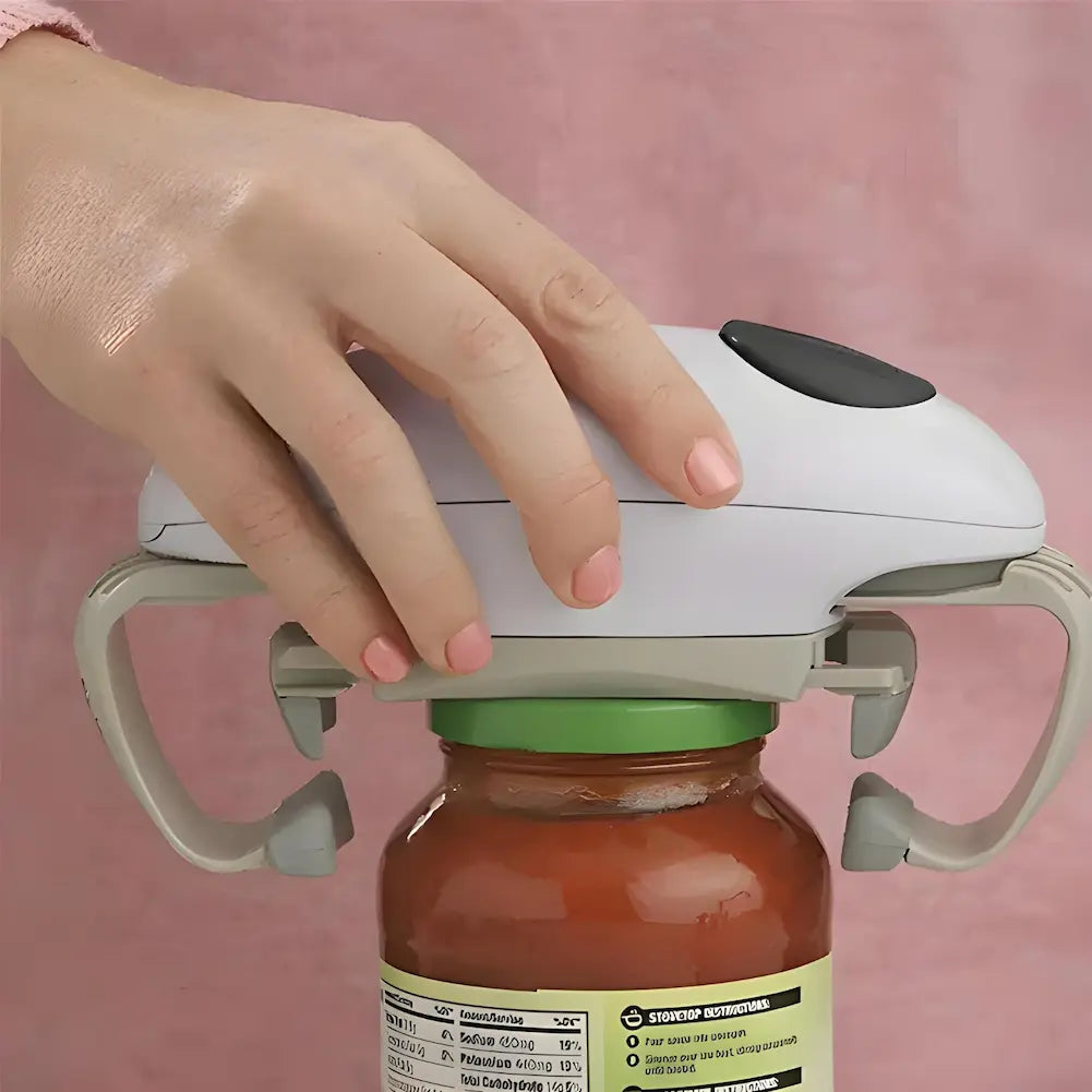 A imagem mostra uma mão usando um abridor de potes automático para abrir um pote de molho com tampa verde. O dispositivo é branco com detalhes cinza e um botão preto no topo. A mão está pressionando o dispositivo sobre a tampa do pote, contra um fundo rosa.